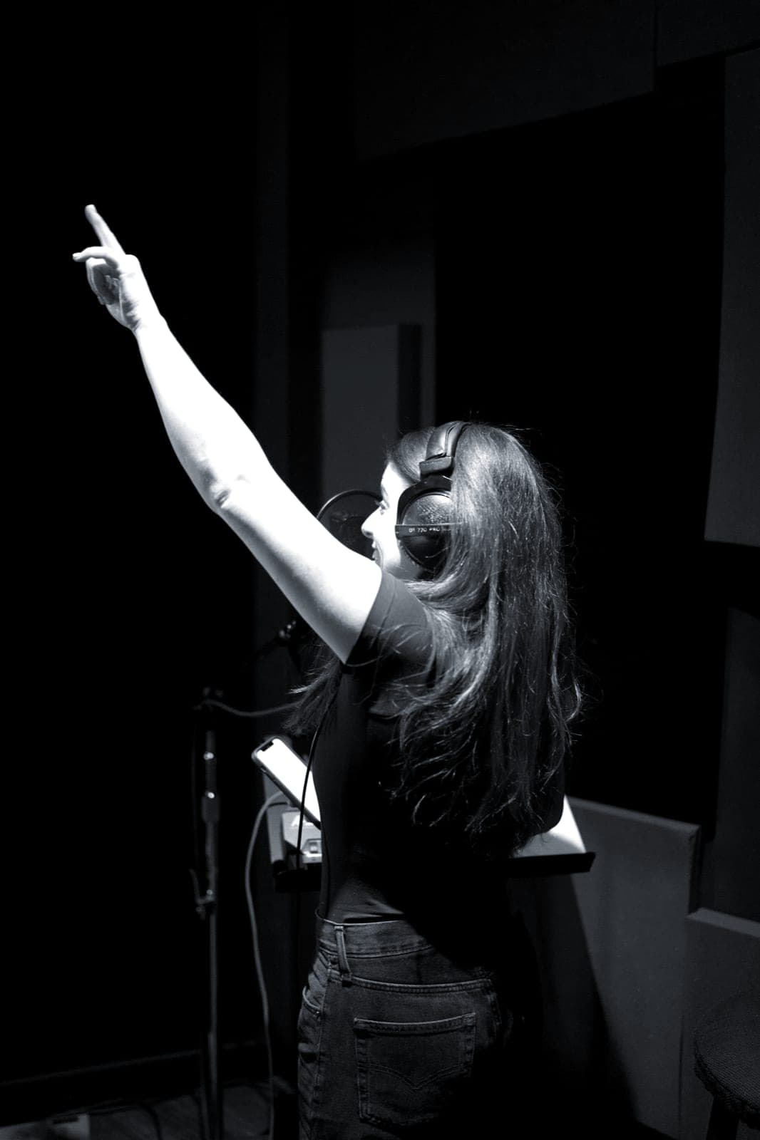 A singer in headphones throws an arm in the air inside the vocal booth.