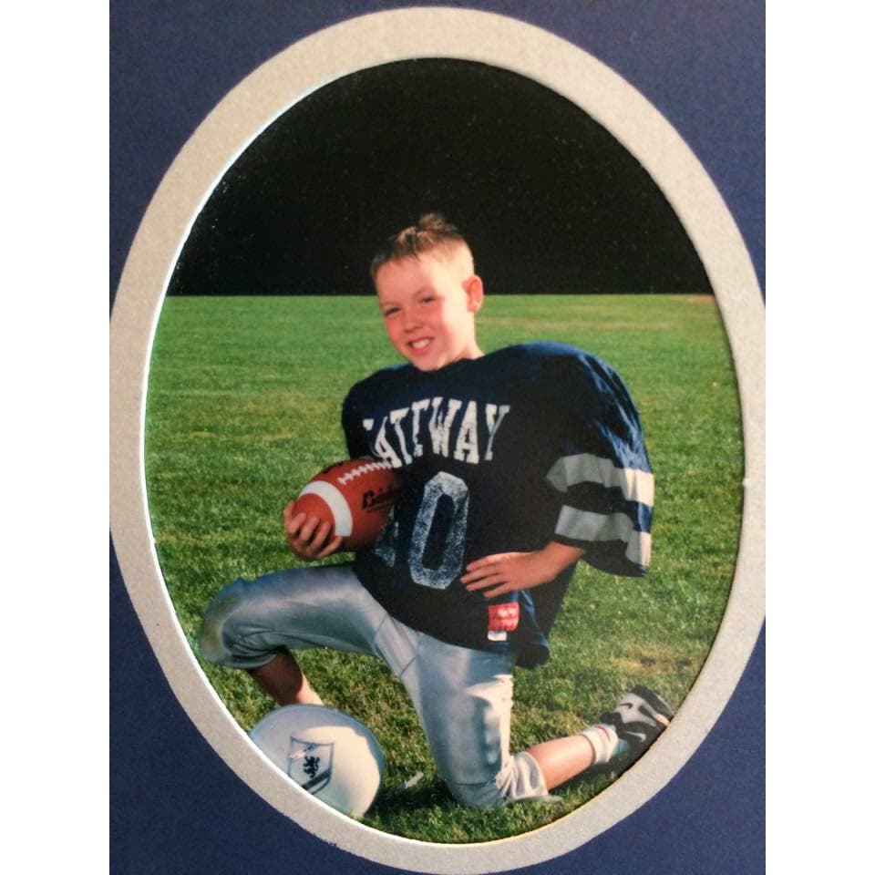 Young Brandon kneeling on the field in his navy Gateway football jersey, holding a football and helmet