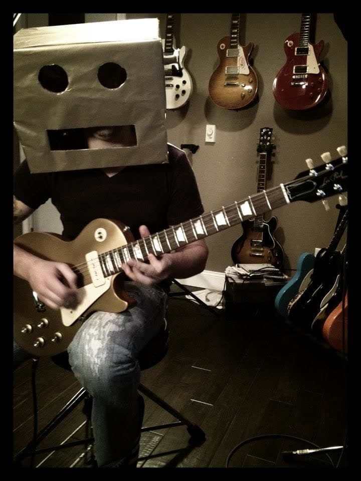 A musician wearing a cardboard robot helmet and playing a guitar in the studio