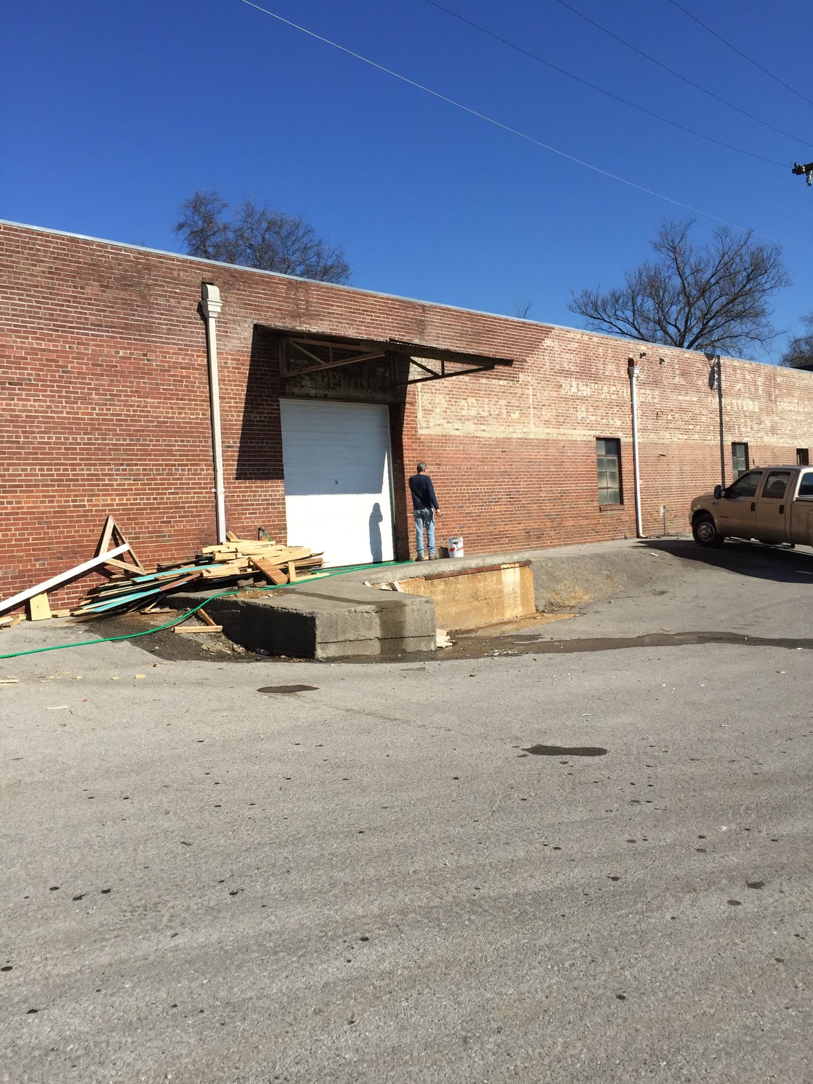 The brick warehouse exterior on Jewel Street: faded ghost signage on the wall, a white garage door, lumber stacked by the loading dock, a work truck parked alongside under blue sky