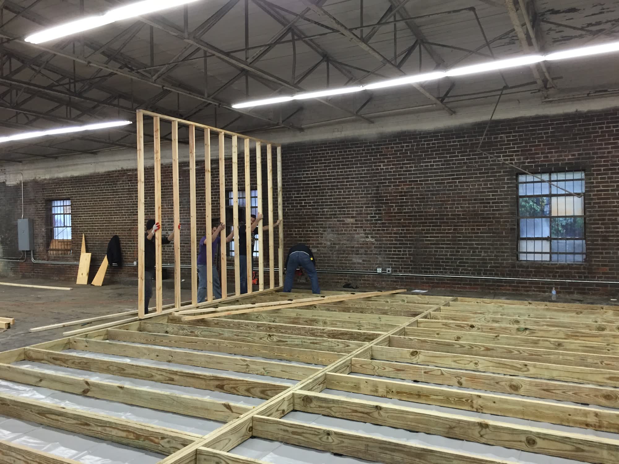 Four friends raising a framed stud wall by hand over fresh floor joists inside the brick warehouse