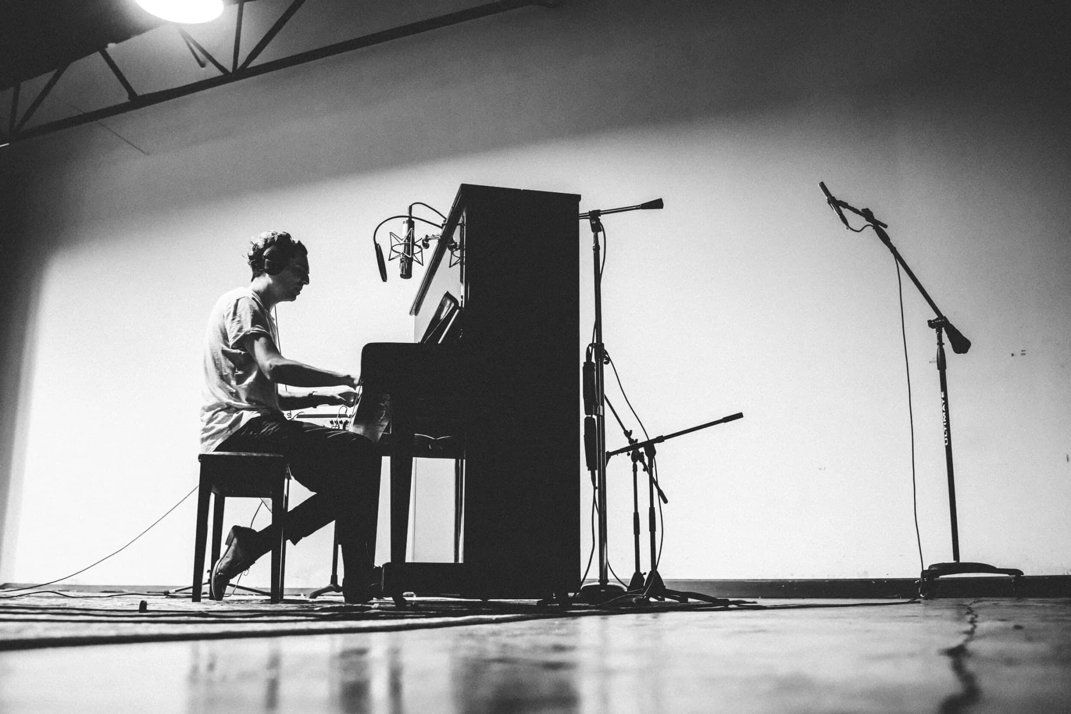 Wide black-and-white photograph of a musician at an upright piano, surrounded by microphones in Studio A