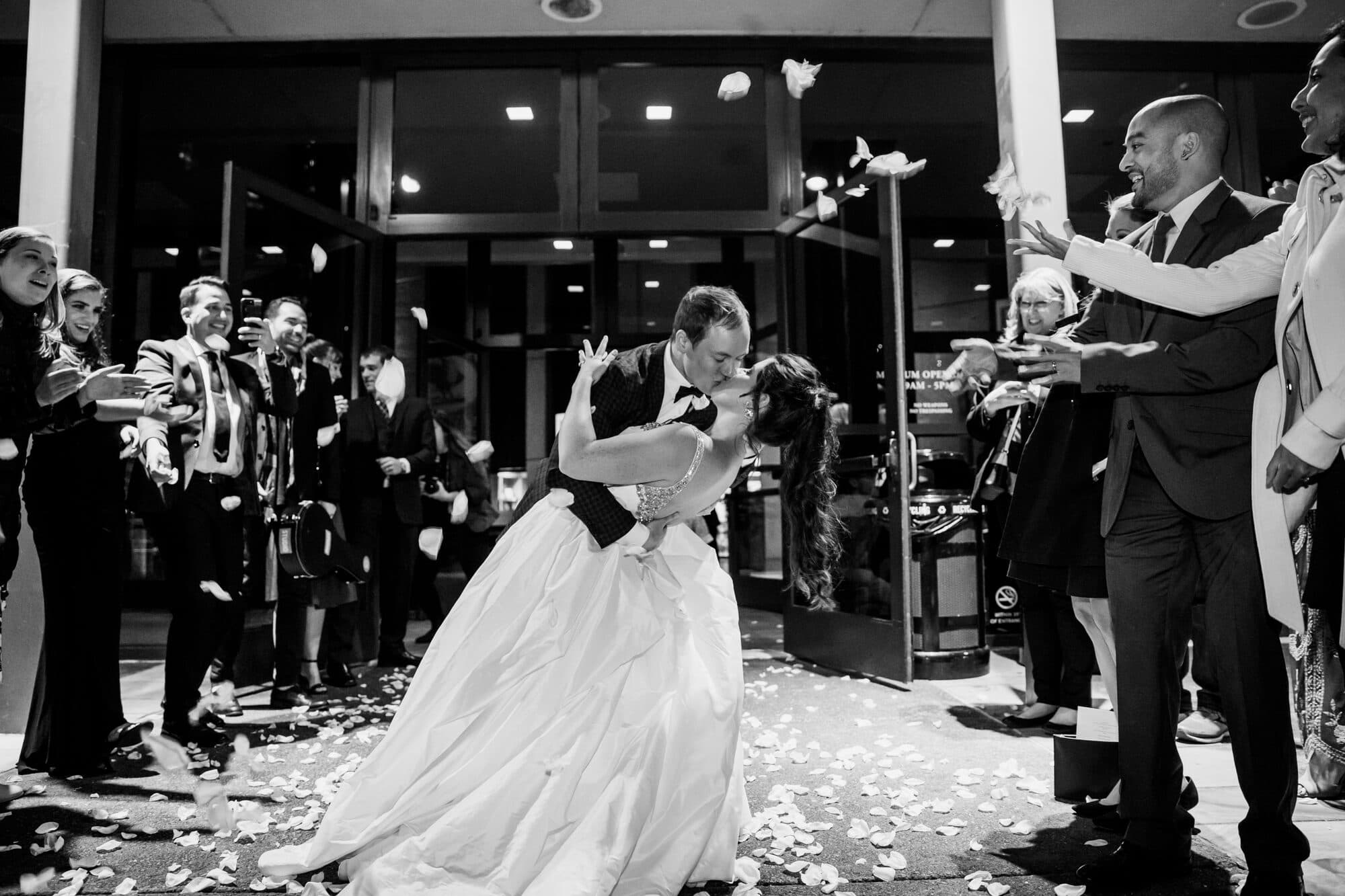 Black-and-white photo of the wedding exit: the groom dips the bride into a kiss as guests toss white rose petals