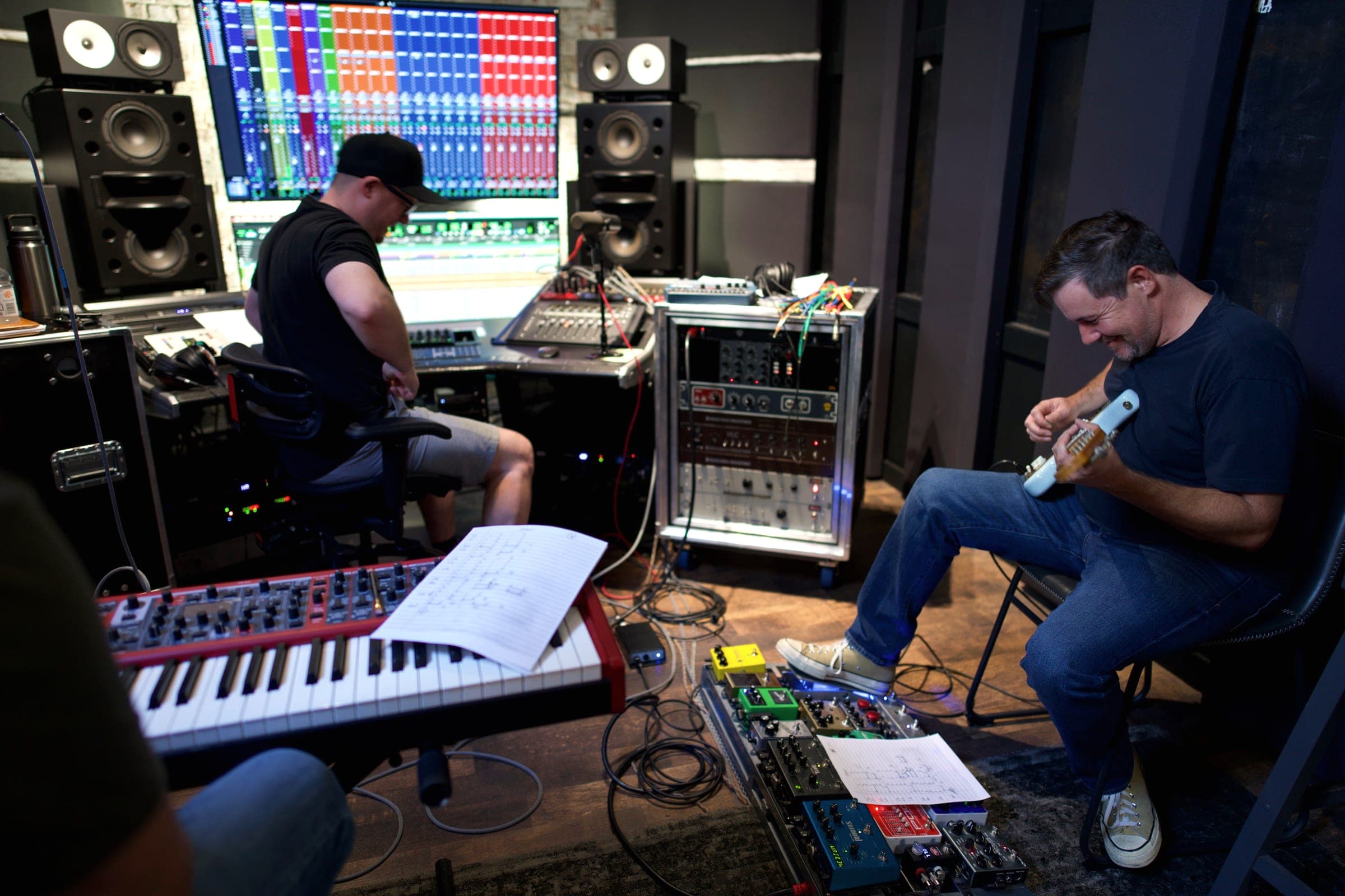 A guitarist with a Jazzmaster, a keys player, and Brandon Metcalf at the desk track together in the studio.